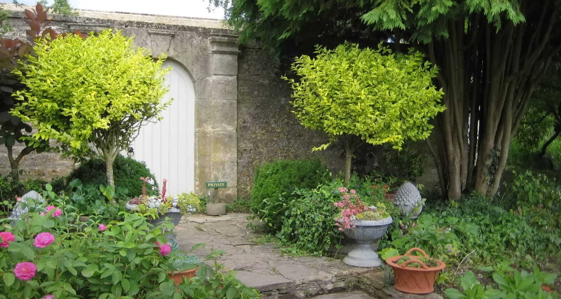 Traditional cottage garden landscape with stone archway entrance, decorative planters, and lush perennial beds